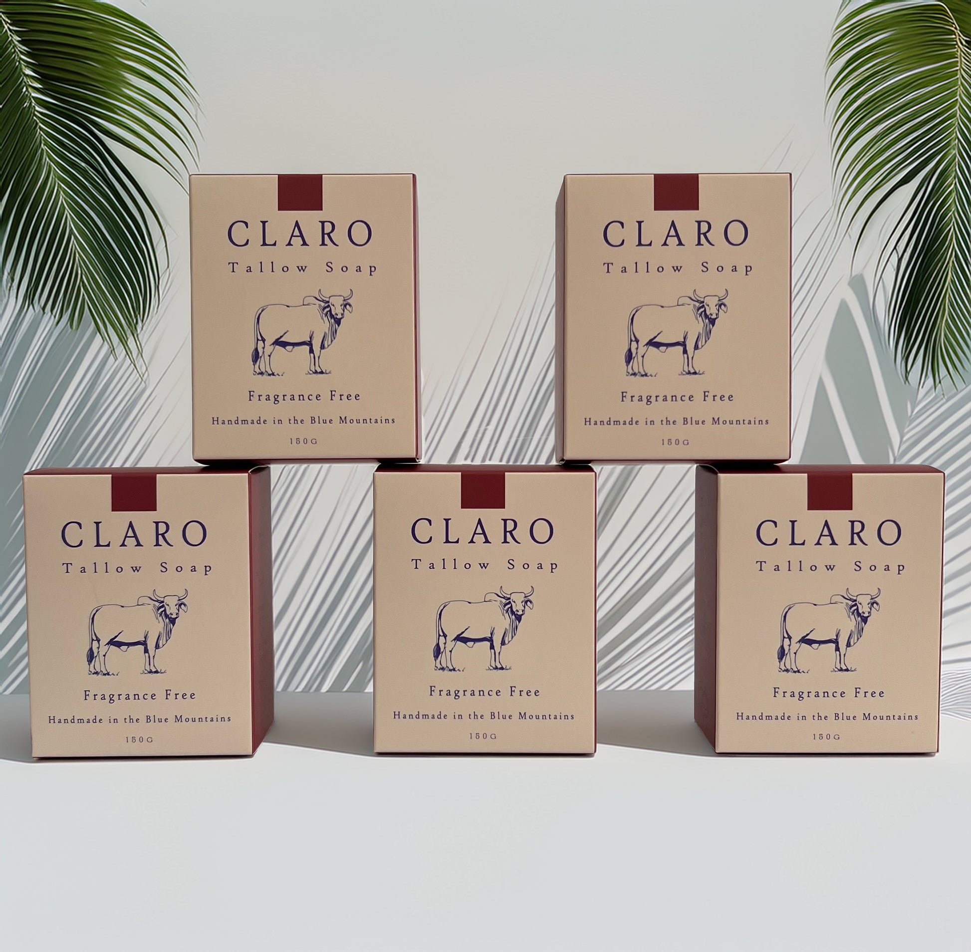 Five boxes of Claro Tallow Soap labelled 'Fragrance Free,' arranged in a pyramid formation against a light background with green palm leaves casting shadows.